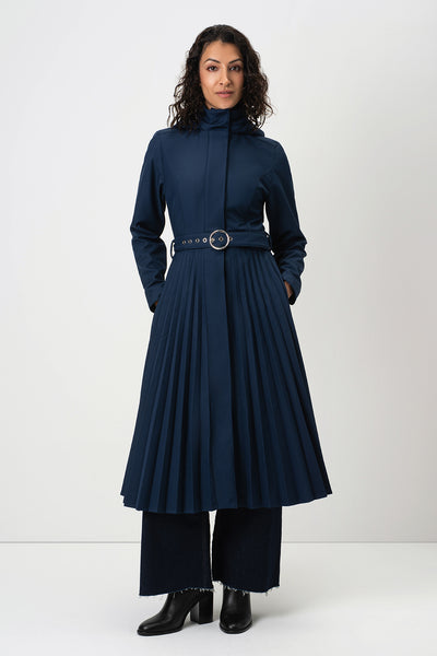 A woman modeling RainSisters dark navy blue long-sleeve trench coat Blue Serenade with a high mock neck, a detachable hood, a thin belt with a round metal buckle, wide-leg trousers, and black ankle boots, set against a plain light gray studio background.