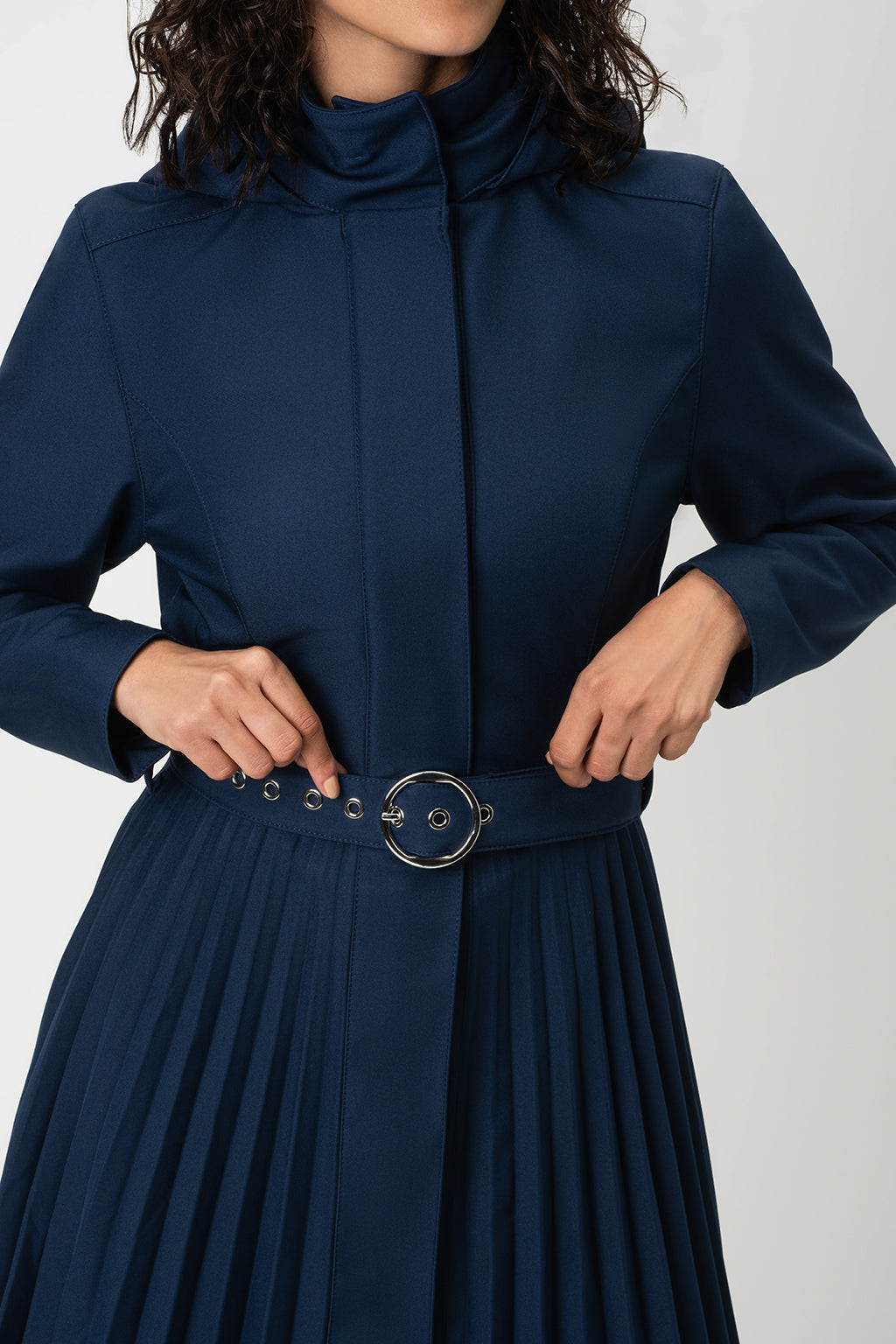 A close-up of a model wearing RainSisters navy blue high-neck trench coat Blue Serenade with a matching pleated skirt and a wide waist belt featuring a large silver-tone circular buckle.