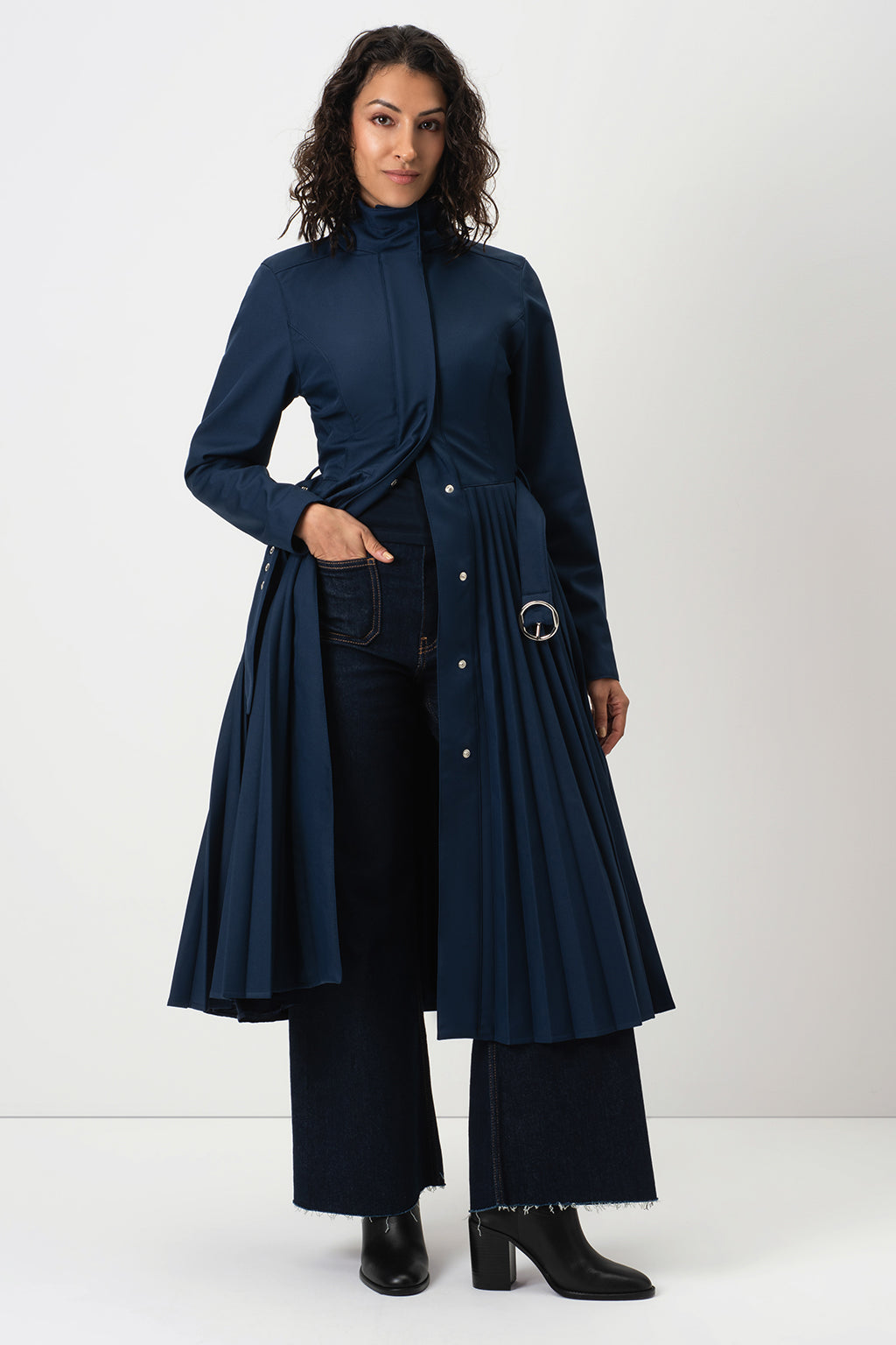 A model wearing RainSisters long navy blue pleated coat Blue Serenade with a high mock neck, paired with dark wide-leg trousers and black ankle boots, photographed against a plain light gray studio background.