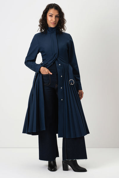 A model wearing RainSisters long navy blue pleated coat Blue Serenade with a high mock neck, paired with dark wide-leg trousers and black ankle boots, photographed against a plain light gray studio background.