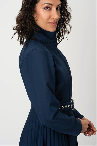 A woman with dark curly hair is shown in profile wearing RainSisters navy blue long-sleeve mock-neck coat Blue Serenade with a pleated skirt and a thin belt with silver grommets, set against a plain light gray background.
