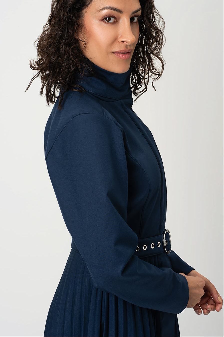 A woman with dark curly hair is shown in profile wearing RainSisters navy blue long-sleeve mock-neck coat Blue Serenade with a pleated skirt and a thin belt with silver grommets, set against a plain light gray background.