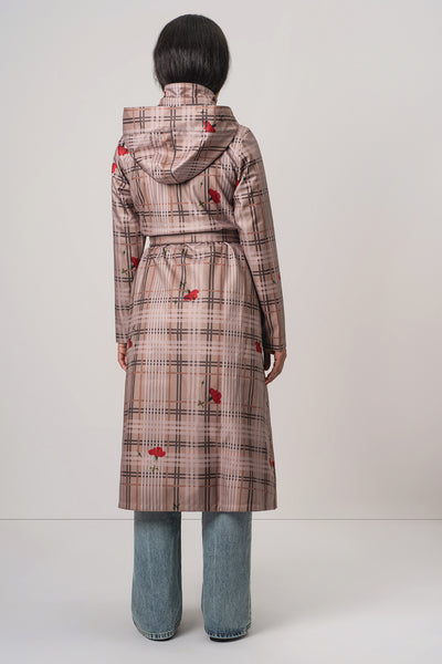 A model viewed from the back wearing RainSisters long hooded coat Blush of the Crown with a beige and brown plaid pattern and scattered red floral accents, paired with wide-leg blue jeans against a plain light gray studio background.
