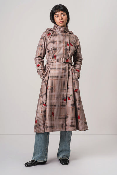 A model wearing RainSisters brown and beige plaid hooded coat Blush of the Crown with red flower motifs, styled with blue jeans and black shoes against a light gray studio background.