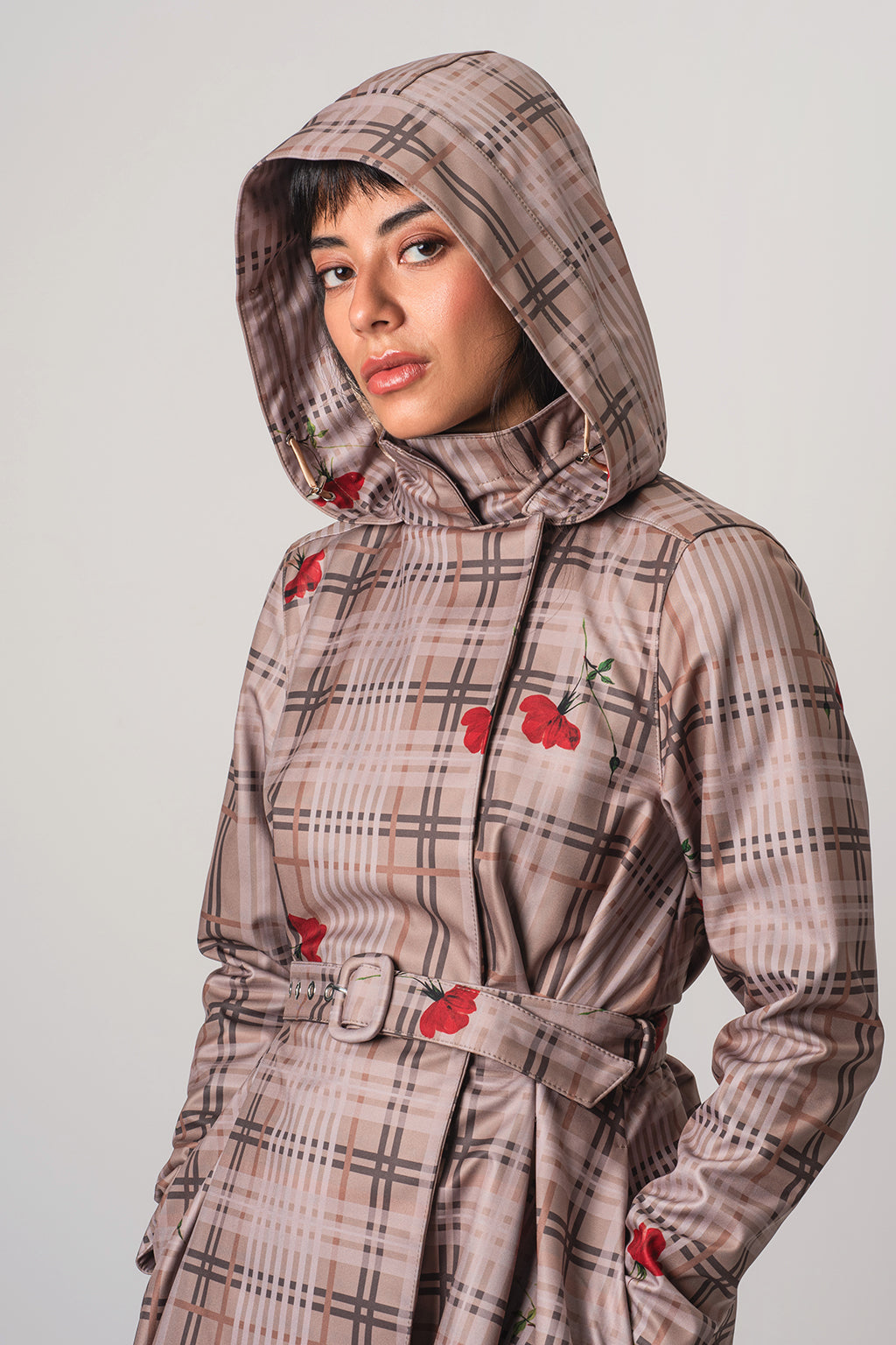 A model wearing RainSisters hooded raincoat Blush of the Crown with a beige and brown plaid pattern and red flower motifs, photographed against a neutral light gray background.