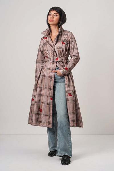 A model wearing RainSisters long, belted trench coat Blush of the Crown in a beige and brown plaid pattern with red rose embroidery, paired with light blue wide-leg jeans and black shoes, against a light gray studio background.