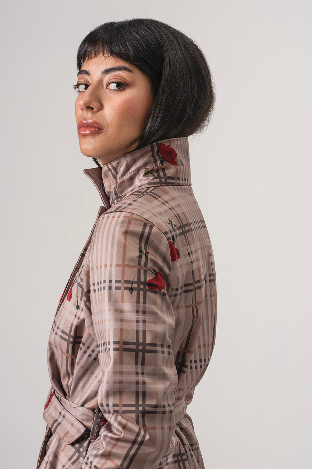 A woman with a short dark hair is shown in profile wearing RainSisters beige plaid coat Blush of the Crown with red floral accents, photographed against a plain light gray studio background.
