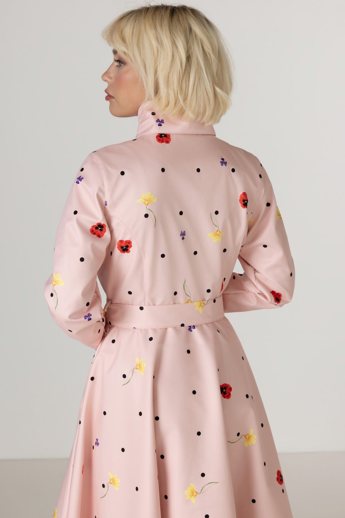 A woman shown from the back wearing RainSisters light pink waterproof coat Cotton Candy with polka dot and colorful flower print, photographed against a plain light gray studio background.