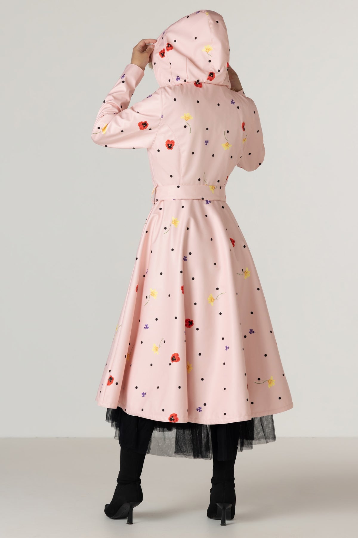 A model viewed from the back wearing RainSisters light pink hooded raincoat Cotton Candy with a colorful floral and polka dot pattern, paired with black tights and black high-heeled boots, against a neutral studio background.