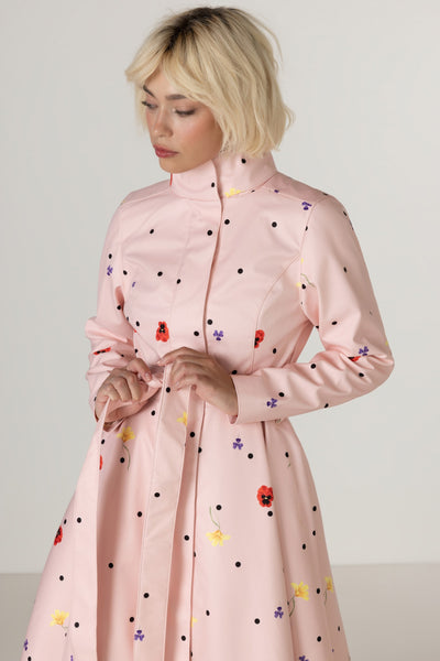A woman wearing RainSisters light pink raincoat Cotton Candy with a high mock neck and a scattered print of black polka dots, red, yellow and purple flowers, shown against a plain light gray studio background.