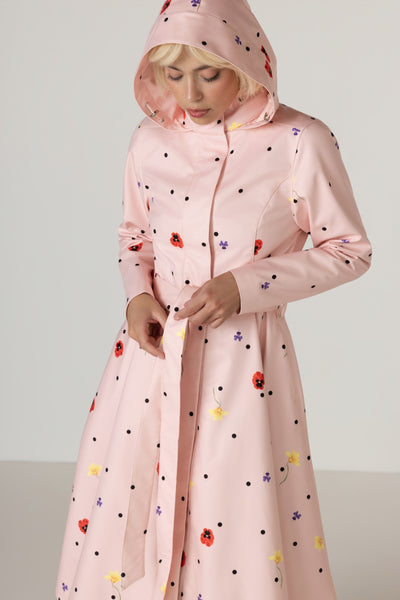A woman wearing RainSisters light pink hooded raincoat Cotton Candy with a multicolor floral and polka dot pattern, shown from the waist up against a plain light gray studio background.