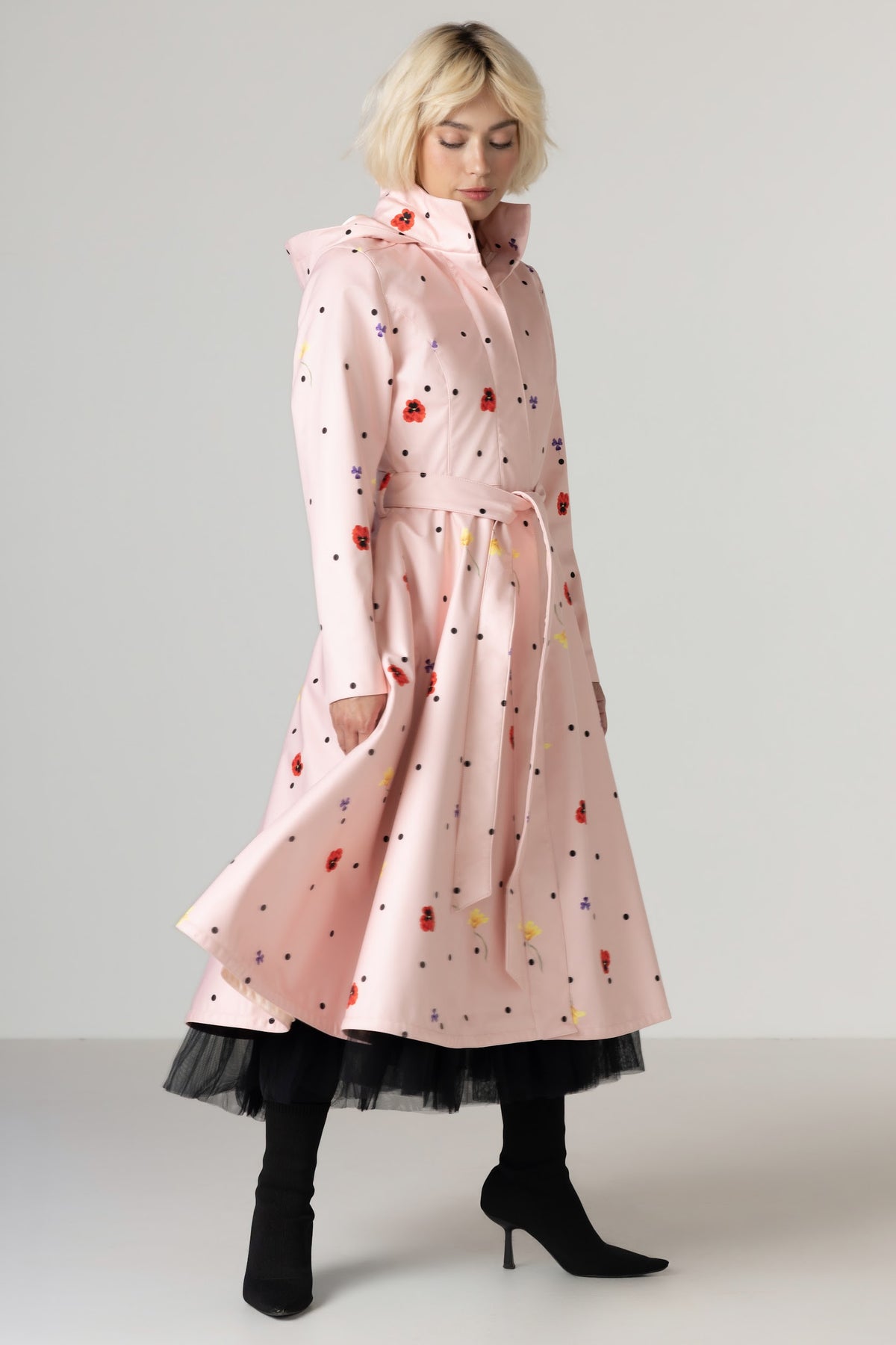 A model wearing RainSisters light pink hooded raincoat Cotton Candy with a colorful scattered print, styled with a black tulle hem and black heeled boots against a neutral studio background.