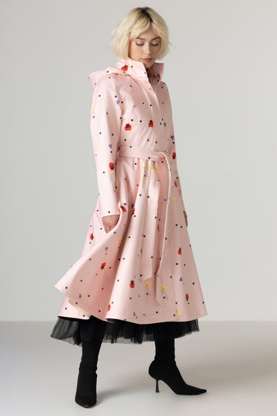 A model wearing RainSisters light pink hooded raincoat Cotton Candy with a colorful scattered print, styled with a black tulle hem and black heeled boots against a neutral studio background.