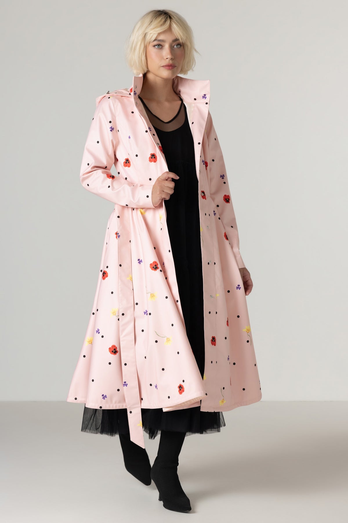 A model wearing RainSisters light pink hooded raincoat Cotton Candy with a colorful retro pattern of dots and small red, yellow, and lilac motifs, styled over a black dress and black boots against a neutral studio background.