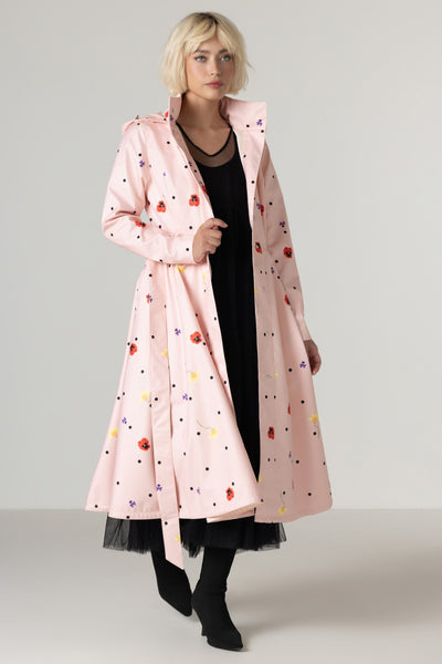 A model wearing RainSisters light pink hooded raincoat Cotton Candy with a colorful retro pattern of dots and small red, yellow, and lilac motifs, styled over a black dress and black boots against a neutral studio background.
