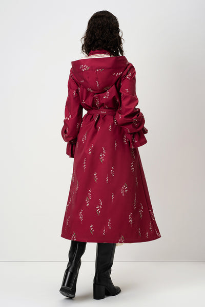 A model viewed from the back wearing RainSisters long burgundy hooded coat Crimson Lily with a white floral pattern, a matching waist tie, and black knee-high boots against a plain white studio background.