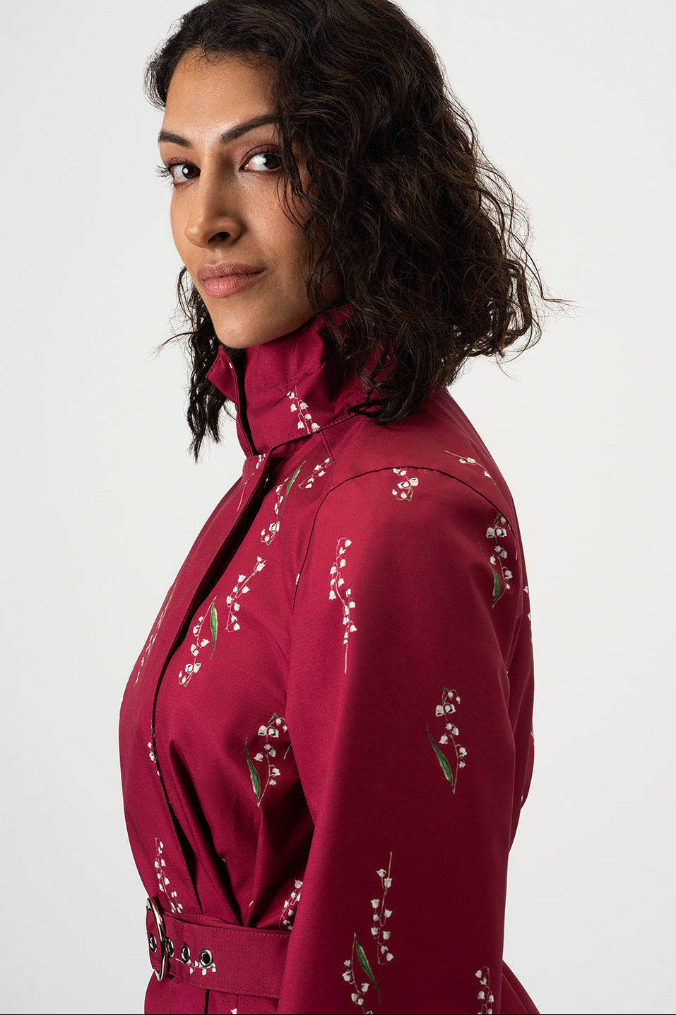 A woman with dark curly hair is shown in profile wearing RainSisters burgundy high-neck jacket Crimson Lily with white floral details, set against a plain light gray background.
