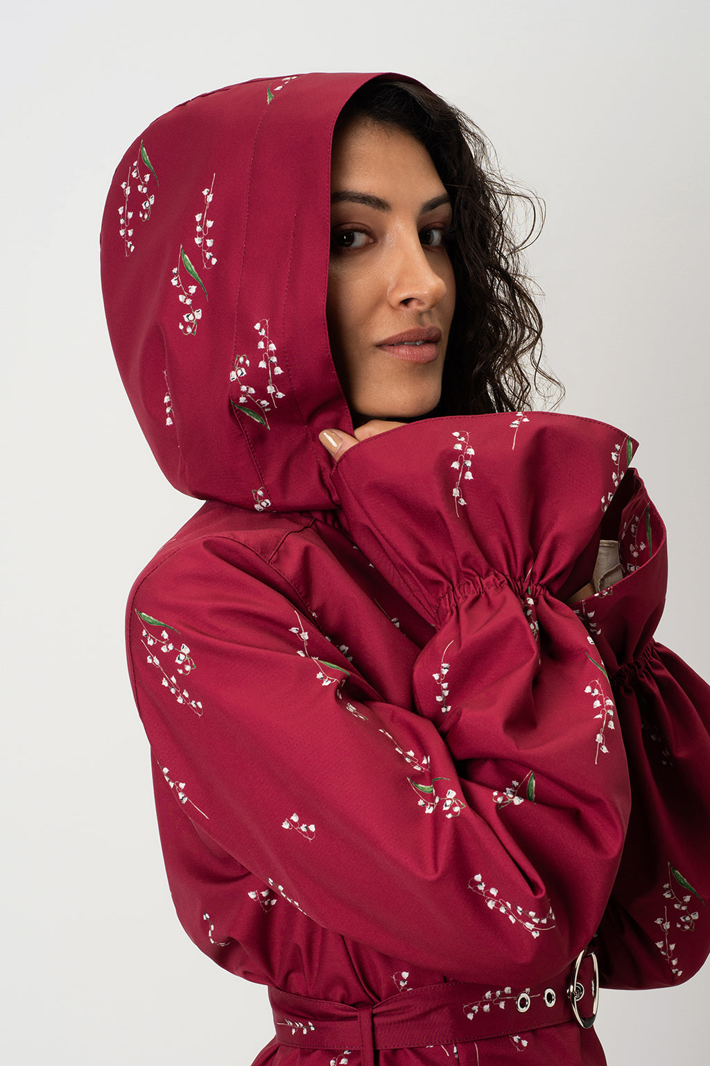 A woman wearing RainSisters deep red hooded waterproof jacket Crimson Lily with white floral print, posed against a light gray studio background.