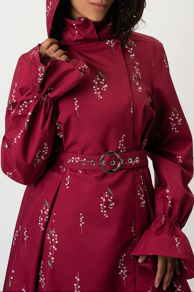 A woman wearing RainSisters hooded burgundy raincoat Crimson Lily with a white floral pattern, a matching belt with silver hardware, and dramatic ruffled cuffs, photographed against a light gray studio background.