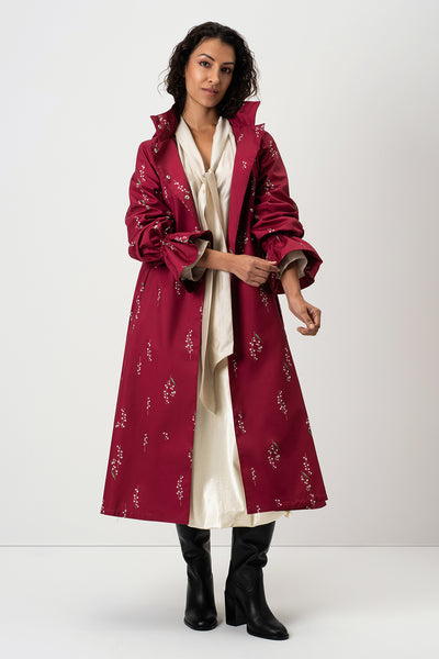 A woman models RainSisters long burgundy trench coat Crimson Lily with small white floral prints, worn over a cream satin set and black knee-high boots against a plain white studio background.