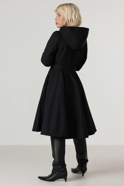 A woman shown from the back wearing RainSisters black hooded coat Deep Emerald with a waist tie, black knee-high boots, and black leggings against a plain light gray studio background.