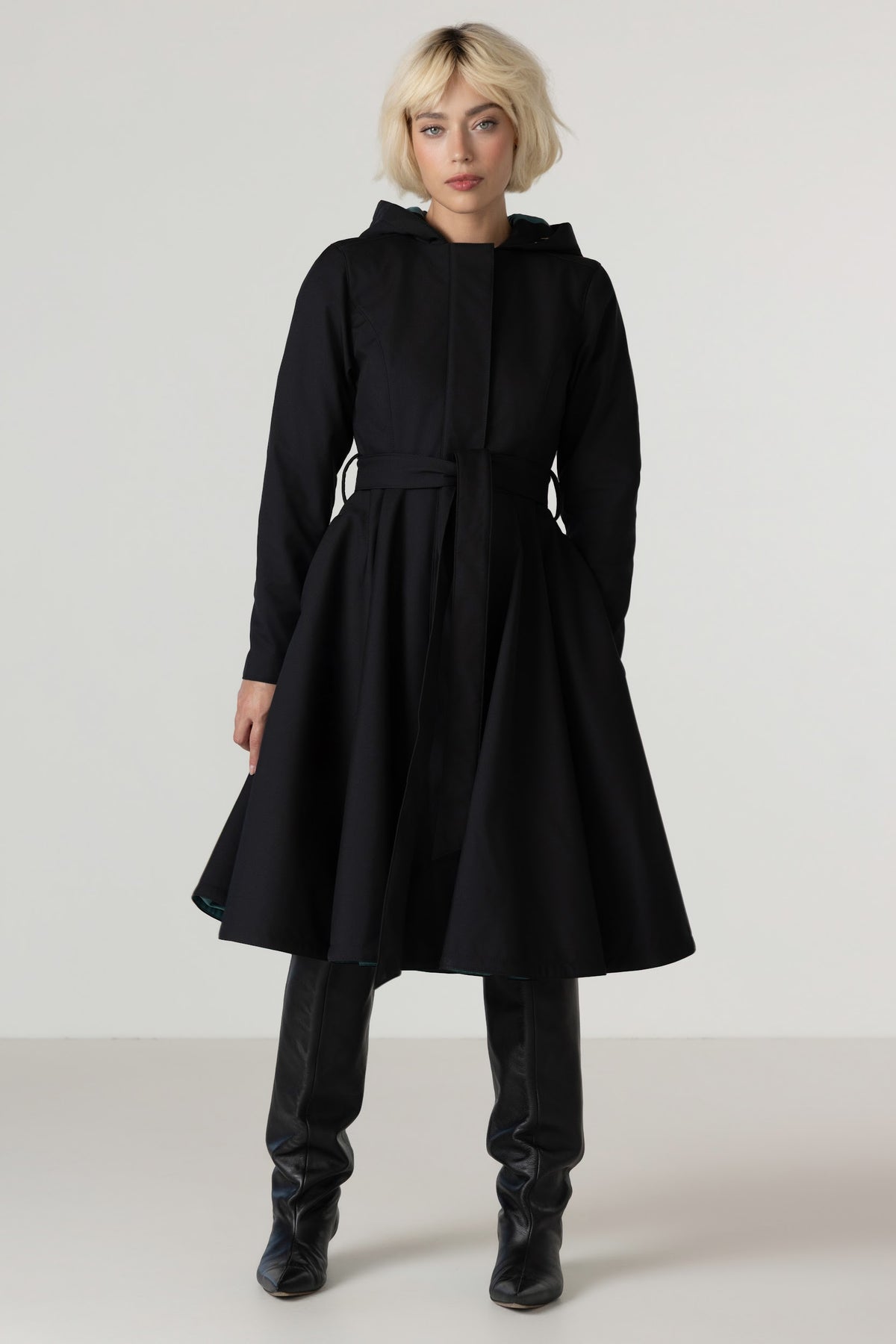 A model with short blonde hair wearing RainSisters black hooded longline coat Deep Emerald with a matching waist tie, black leather-look boots, and black pants, standing against a light gray studio background.