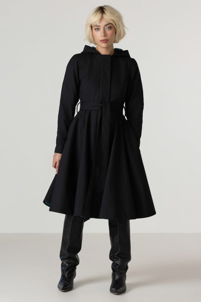 A model with short blonde hair wearing RainSisters black hooded longline coat Deep Emerald with a matching waist tie, black leather-look boots, and black pants, standing against a light gray studio background.