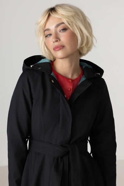 A blonde woman wearing RainSisters black hooded trench coat Deep Emerald with a matching waist tie and emerald green lining, layered over a red top, photographed against a plain light gray studio background.