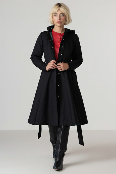 A model with short blonde hair wearing RainSisters long black hooded coat Deep Emerald over a red top and black pants, standing against a light gray studio background.