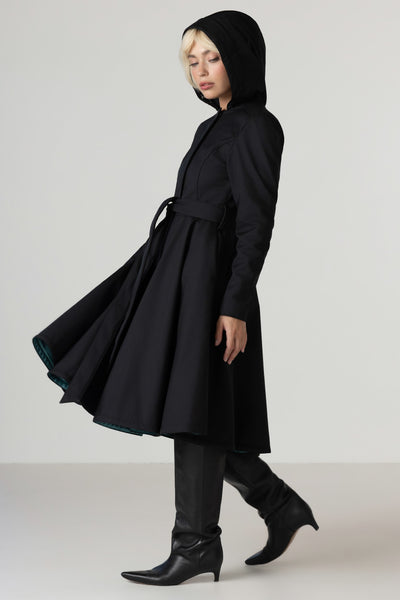 A model wearing RainSisters black hooded longline coat Deep Emerald with a matching belt, paired with black pointed-toe heeled boots, photographed against a plain light gray studio background.