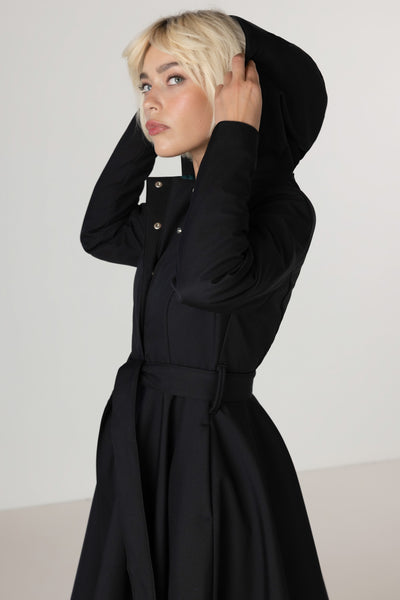 A model with short blonde hair wearing RainSisters black hooded long-sleeve waterproof coat Deep Emerald with a belted waist, posing against a neutral light gray background.