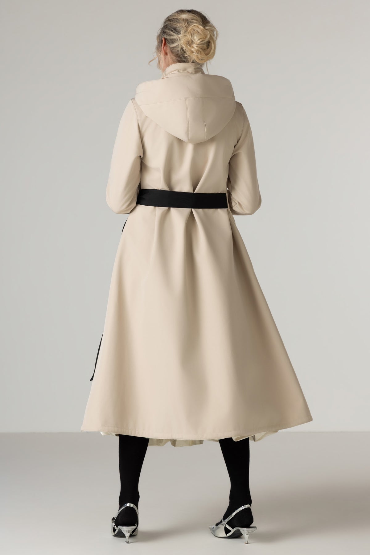 A woman shown from the back wearing RainSisters long cream-colored hooded coat Desirable Bow with a black waist belt, black tights, and silver pointed shoes against a light gray studio background.