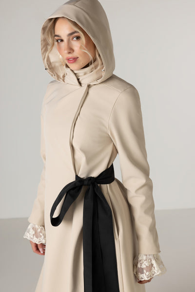 A woman wearing RainSisters beige hooded longline coat Desirable Bow with a black waist tie styled with lace ruffled cuffs, photographed against a plain light gray studio background.
