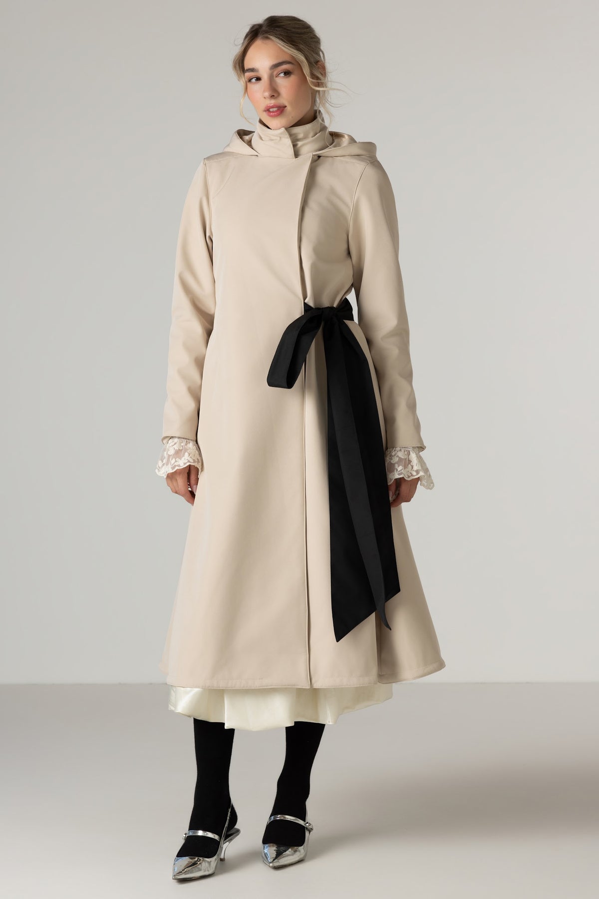 A woman models RainSisters long beige hooded coat with a black waist tie Desirable Bow, worn over a white dress. She is also wearing black tights and silver pointed shoes against a neutral light gray studio background.