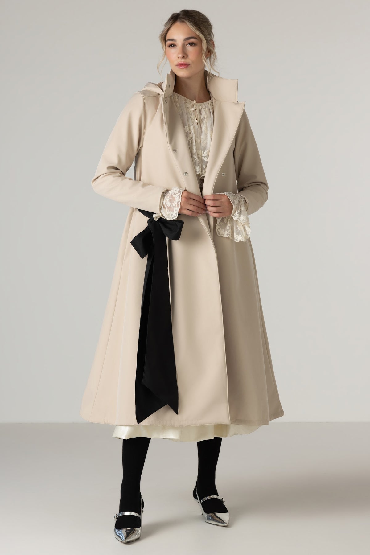 A woman models RainSisters long beige trench coat Desirable Bow with a large black sash tied at the waist, worn over a cream lace-trimmed blouse and paired with black tights and metallic shoes, against a light gray studio background.