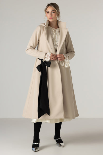 A woman models RainSisters long beige trench coat Desirable Bow with a large black sash tied at the waist, worn over a cream lace-trimmed blouse and paired with black tights and metallic shoes, against a light gray studio background.