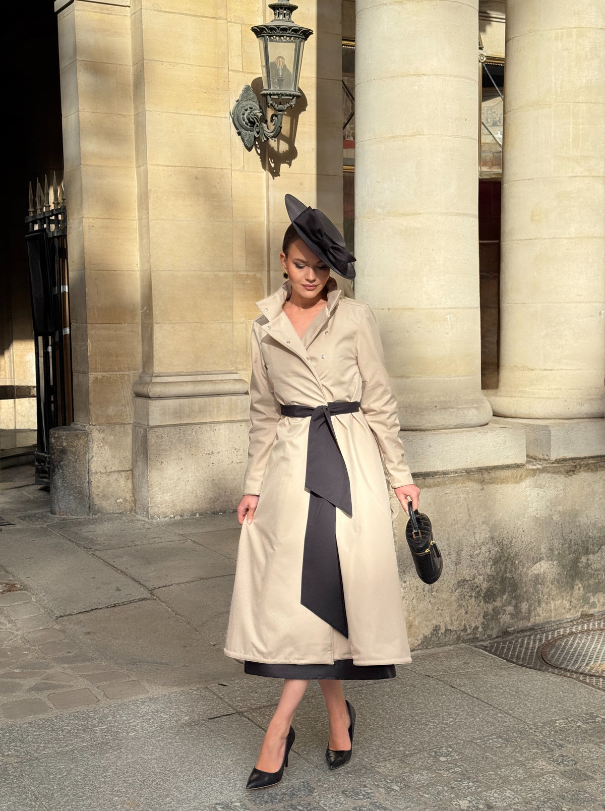 A woman stands on a stone sidewalk in front of a classical building with large columns, wearing RainSisters beige trench coat Desirable Bow with a black sash and matching dark hat, holding a small dark handbag.