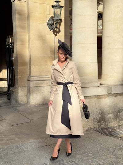 A woman stands on a stone sidewalk in front of a classical building with large columns, wearing RainSisters beige trench coat Desirable Bow with a black sash and matching dark hat, holding a small dark handbag.