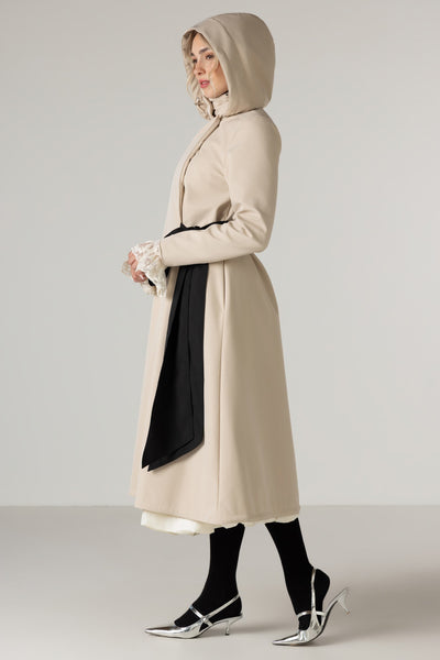 A woman modeling RainSisters long beige hooded coat Desirable Bow with a black draped tie styled white lace cuffs, black tights and metallic pointed heels, against a light gray studio background.