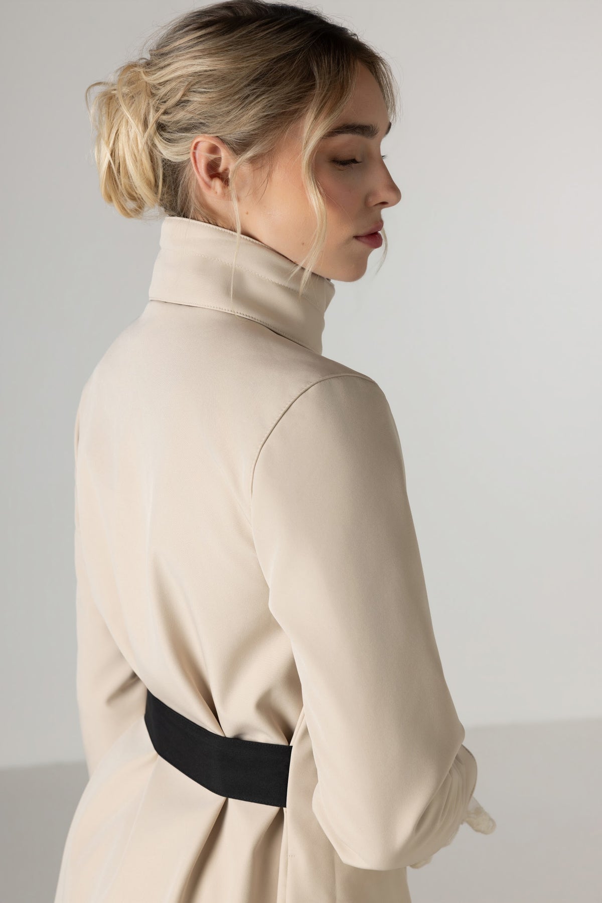 A woman shown from the back wearing RainSisters light beige coat Desirable Bow with detached hood and  wide black waist belt, set against a plain light gray studio background.