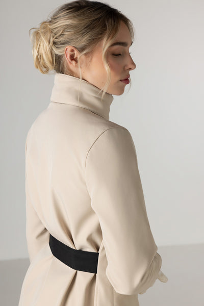 A woman shown from the back wearing RainSisters light beige coat Desirable Bow with detached hood and  wide black waist belt, set against a plain light gray studio background.