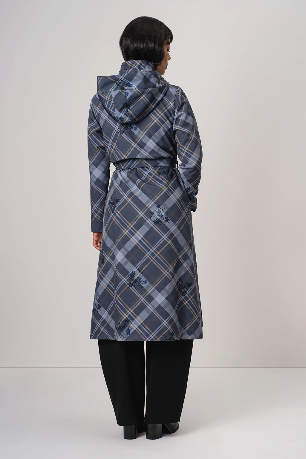 A woman viewed from the back wearing RainSisters long hooded coat Dutchess in Blue with a blue, gray, and white plaid pattern and black trousers, standing against a plain light gray studio background.