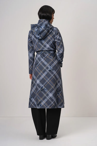 A woman viewed from the back wearing RainSisters long hooded coat Dutchess in Blue with a blue, gray, and white plaid pattern and black trousers, standing against a plain light gray studio background.