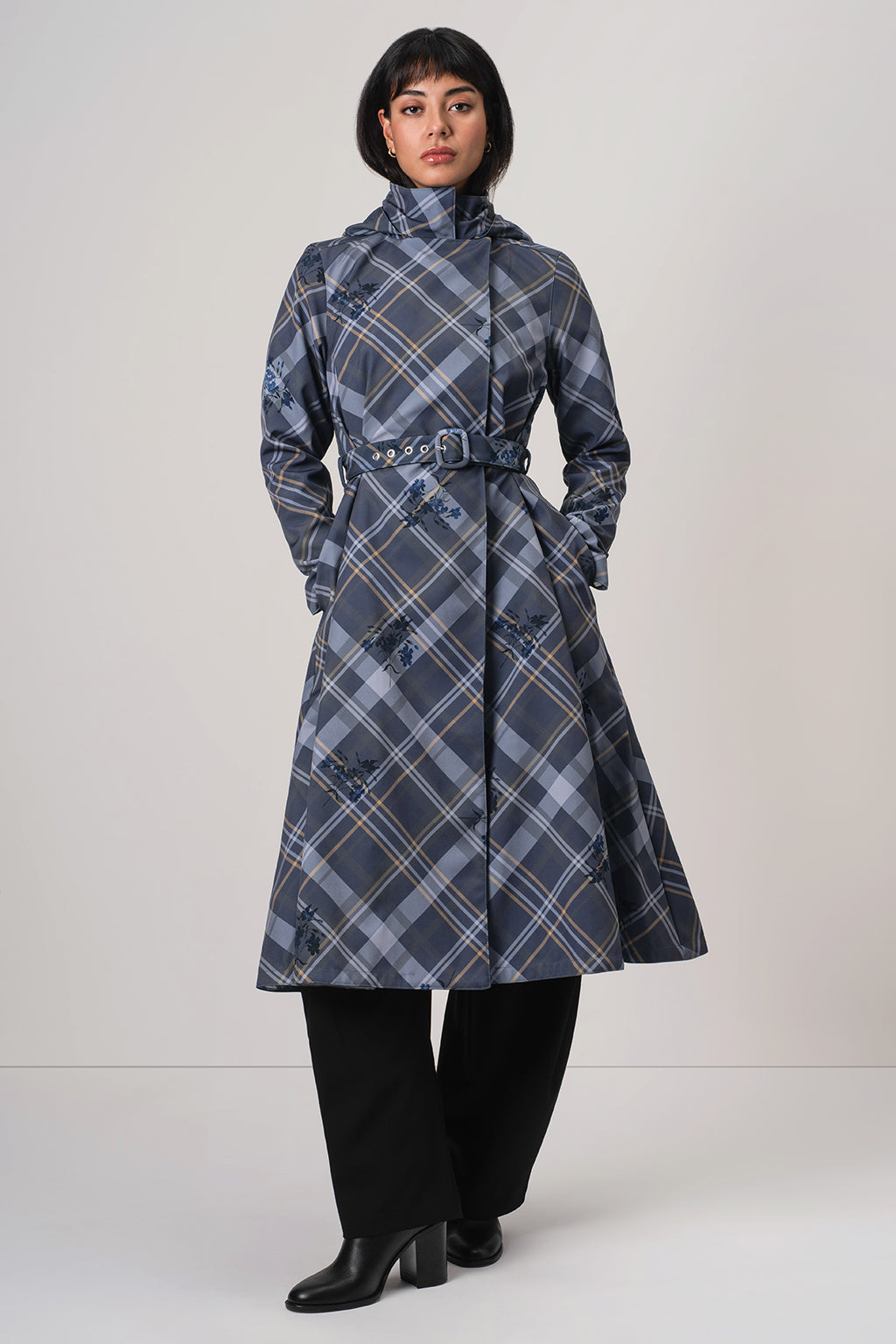 A model wearing RainSisters mid-length blue plaid coat Dutchess in Blue with a belted waist, black wide-leg trousers, and black boots, standing against a light gray studio background.