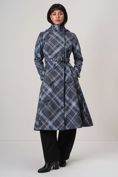 A model wearing RainSisters mid-length blue plaid coat Dutchess in Blue with a belted waist, black wide-leg trousers, and black boots, standing against a light gray studio background.