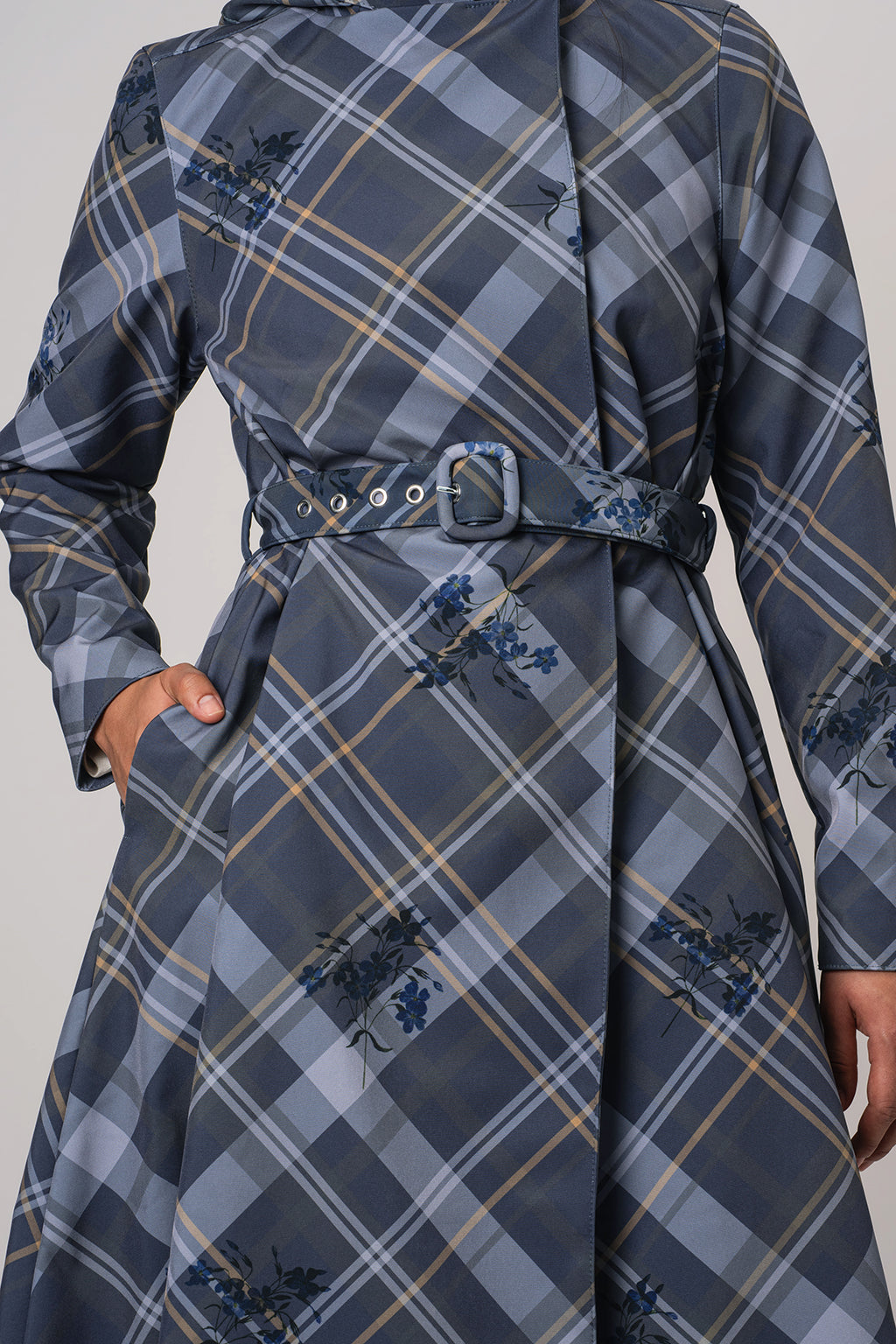 A close-up of a person wearing RainSisters long-sleeve belted coat Dutchess in Blue with a gray plaid pattern and small blue floral motifs, shown against a plain light gray background.