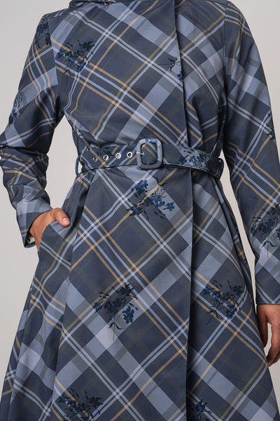 A close-up of a person wearing RainSisters long-sleeve belted coat Dutchess in Blue with a gray plaid pattern and small blue floral motifs, shown against a plain light gray background.