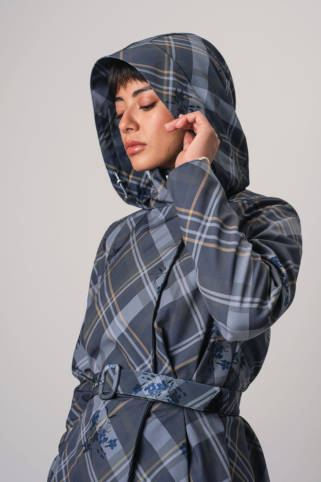 A model wearing RainSisters hooded raincoat Dutchess in Blue with a blue, gray, and beige plaid pattern, featuring a matching waist belt, set against a plain light gray background.