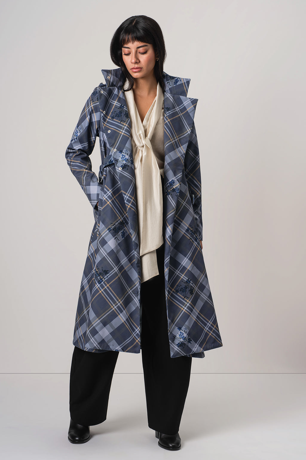 A model wearing RainSisters long blue and gray plaid coat Dutchess in Blue over a cream blouse, black trousers, and black shoes, standing against a light gray studio background.