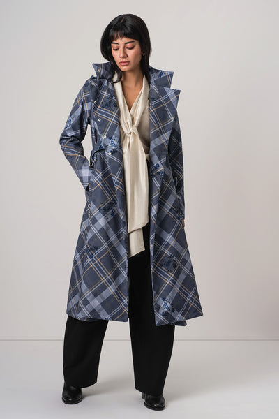 A model wearing RainSisters long blue and gray plaid coat Dutchess in Blue over a cream blouse, black trousers, and black shoes, standing against a light gray studio background.